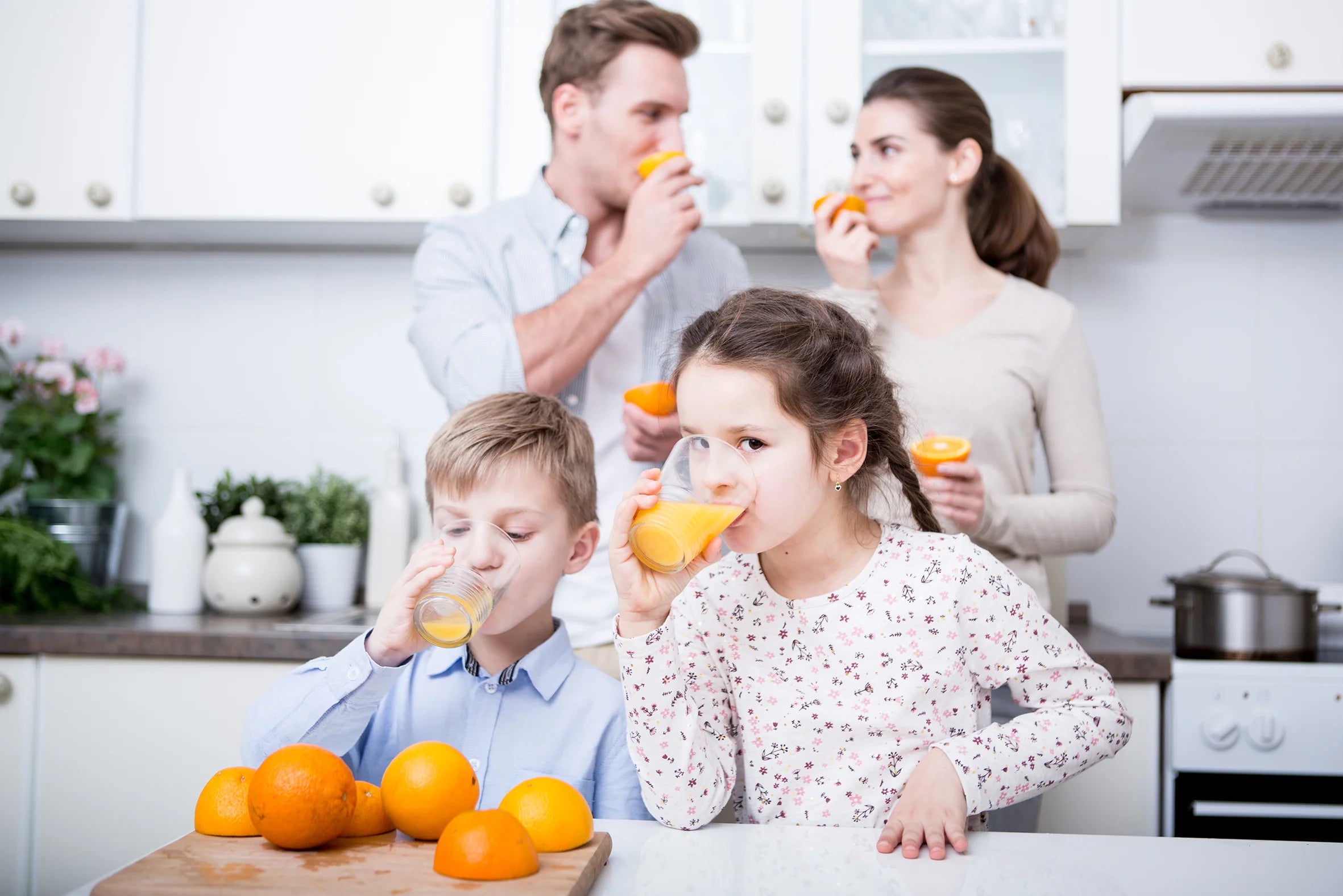 Eye-level, medium shot of a family of four enjoying fresh citrus juice in a bright white kitchen, with a young boy and a girl with braided hair in the foreground drinking from glasses next to whole and cut oranges on a wooden board, while their parents stand out of focus in the background tasting orange halves.