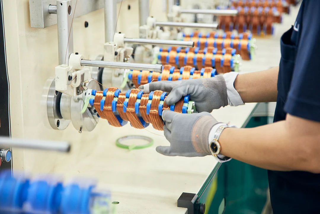 Side eye-level shot of a worker in a dark blue shirt and grey protective gloves working at an automated factory assembly line station, manually adjusting copper induction motor coil windings wrapped around a blue and metal core component for a Kuvings slow juicer, with more motor components visible along the machinery in the background.