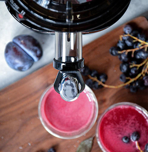 High-angle overhead close-up shot of a Kuvings slow juicer spout with a chrome-tipped smart cap positioned above a glass of fresh purple juice, arranged on a wooden cutting board next to whole plums and dark grapes.