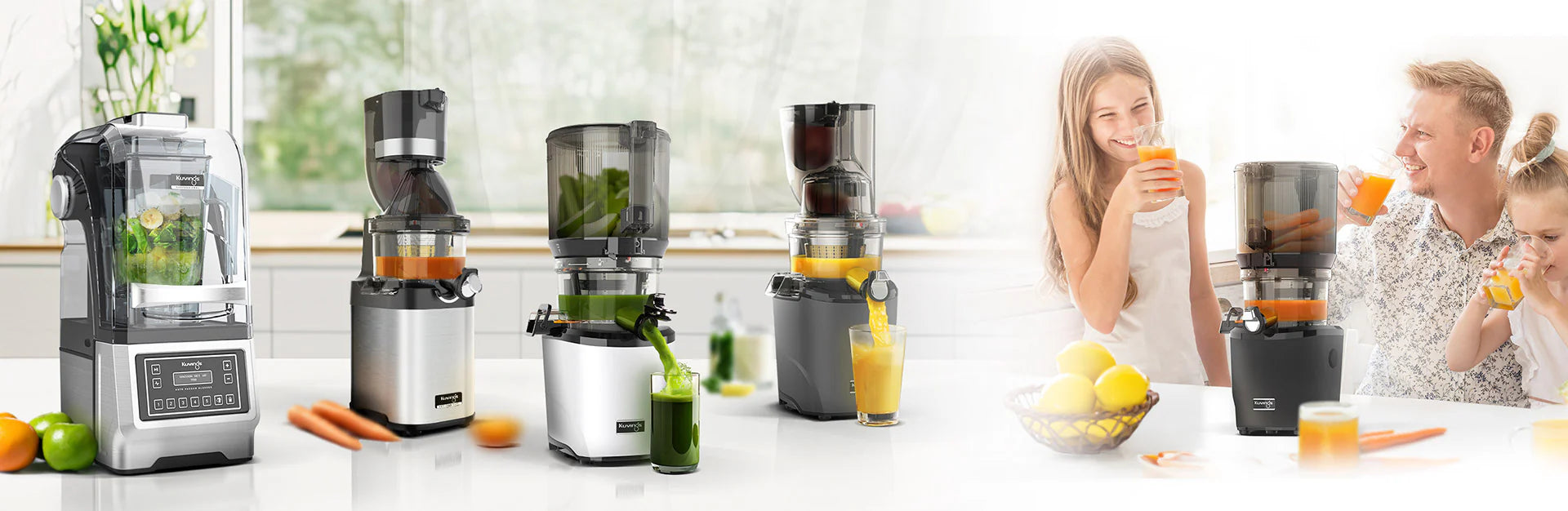 Wide eye-level horizontal marketing banner showing a lineup of four different Kuvings kitchen appliances on a bright kitchen countertop, including a professional vacuum blender and three distinct whole slow juicer models processing various juices, transitioned on the right into a bright lifestyle scene of a happy father and his two young daughters smiling and drinking freshly squeezed orange juice.