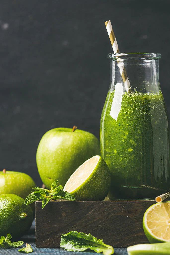Glass bottle of green smoothie with a straw, surrounded by green apples and limes on a dark background