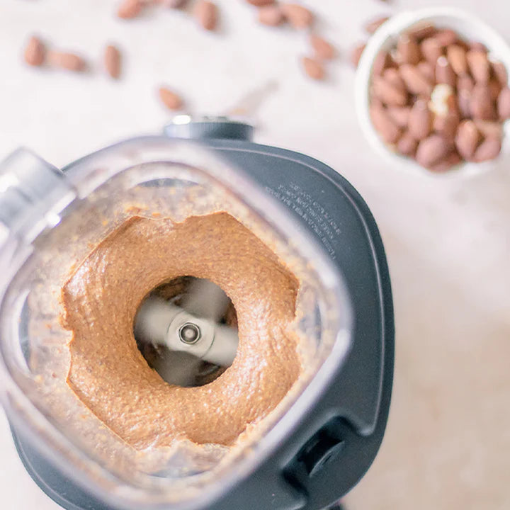Hochwinkelige Draufsicht von oben in den geöffneten Behälter eines Kuvings SV-500 Vakuum-Mixers, während er dicke, cremige Mandelbutter um seine rotierenden Klingen verarbeitet, mit ganzen Mandeln, die im Hintergrund auf der hellen Arbeitsplatte verstreut sind.