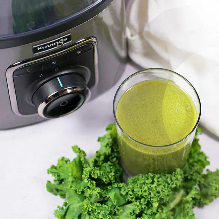 Hochwinkelige Nahaufnahme der silbernen Motorbasis und des Steuerknopfs eines Kuvings SV-500ME Vakuum-Mixers neben einem klaren Glas, gefüllt mit einem lebendigen grünen Smoothie, der auf einem Bett aus frischen Grünkohlblättern ruht.