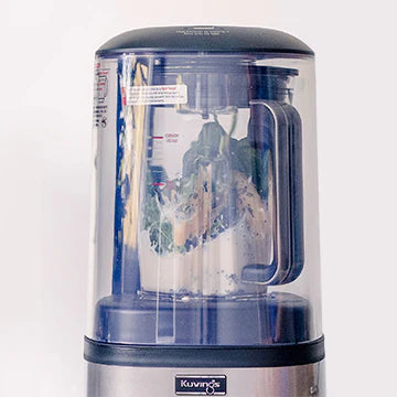 Frontale Nahaufnahme auf Augenhöhe des oberen Teils eines Kuvings SV-500S Vakuum-Mixers im geschlossenen, durchsichtigen Schallschutz, der grünes Blattgemüse und Milch im Mixbehälter vor einem hellen Hintergrund zeigt.