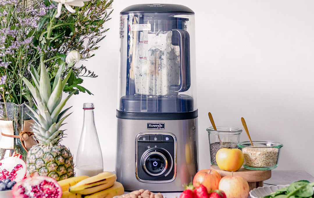 Frontale Aufnahme auf Augenhöhe des Kuvings SV-500S Vakuum-Standmixers auf einer Küchenarbeitsplatte, der eine cremige Mischung mixt, umgeben von einer üppigen Auswahl an frischem Obst, darunter eine Ananas, Bananen, geschnittene Granatäpfel und Äpfel, sowie Gläser mit Samen und Getreide.