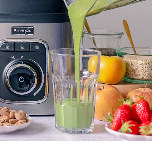 Nahaufnahme auf Augenhöhe eines lebendigen grünen Smoothies, der aus dem Behälter eines Kuvings SV-500S Vakuum-Mixers in ein hohes klares Glas gegossen wird, positioniert neben dem silbernen Bedienfeld des Mixers mit frischen Erdbeeren, Äpfeln und Mandeln auf der Theke.