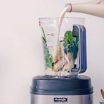 Frontale Ansicht auf Augenhöhe eines Kuvings SV-500S Vakuum-Mixer-Behälters, während Pflanzenmilch aus einer Glasflasche über frisches grünes Blattgemüse und Fruchtstücke im Behälter gegossen wird, vor einem hellen Hintergrund.