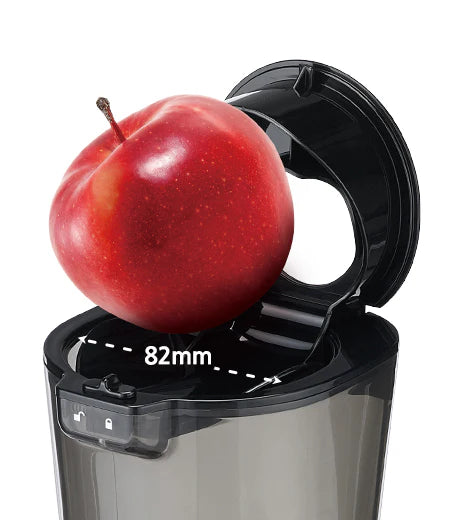 High-angle close-up studio shot showing a large red apple sitting inside the wide-mouth flip-gate feeding chute of a Kuvings whole slow juicer, accented by a white double-sided measurement arrow indicating an 82mm diameter against a white background.