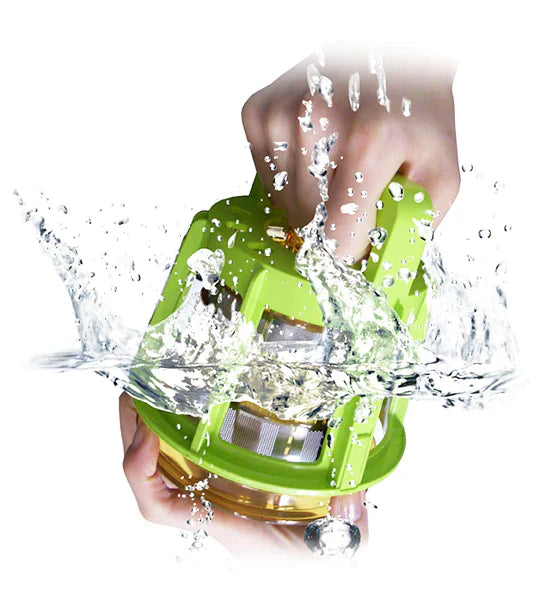 Close-up studio shot of hands using the green rotary cleaning tool to wash a Kuvings juicer metal mesh strainer, submerged under splashing water that creates dynamic bubbles and droplets against a white background.