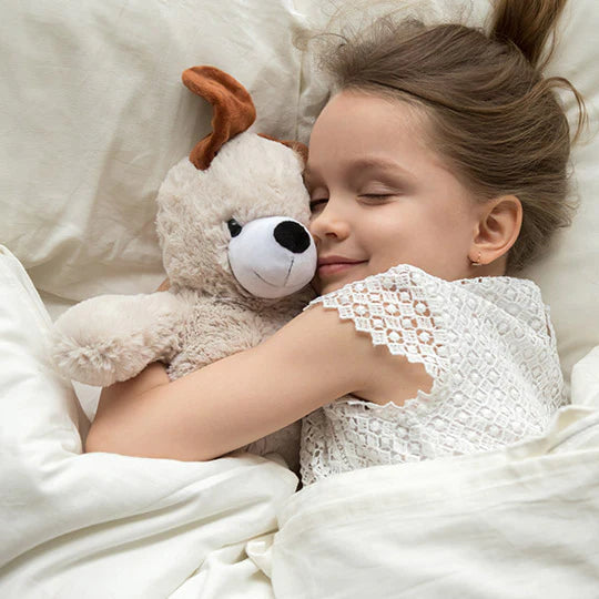 Close-up lifestyle shot of a young girl with closed eyes smiling peacefully while sleeping in a cozy bed with white linens, tightly hugging a fluffy plush stuffed toy dog with brown ears.