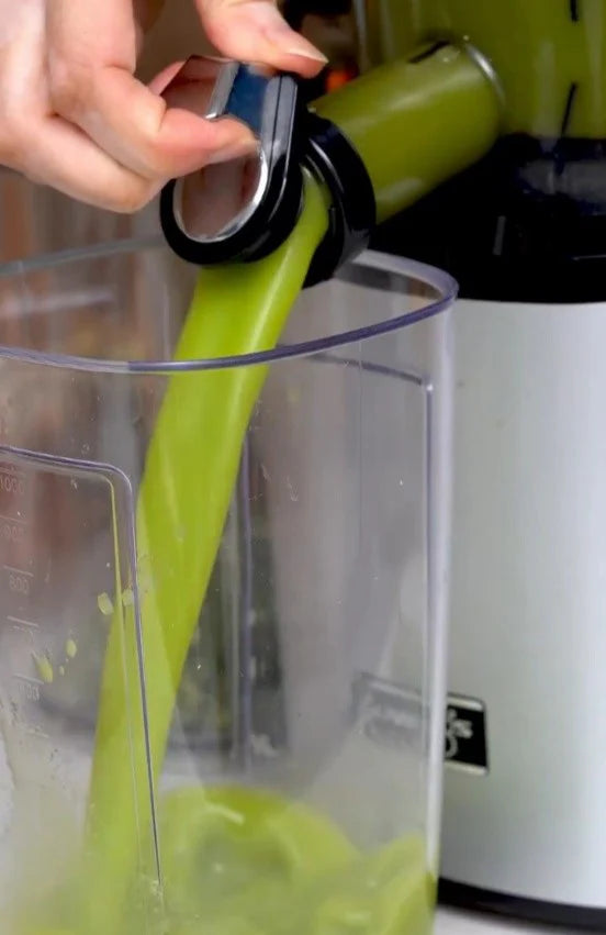 Kuvings green smoothie held by a woman in a kitchen with fresh fruits and vegetables on the counter for healthy homemade smoothie recipes.